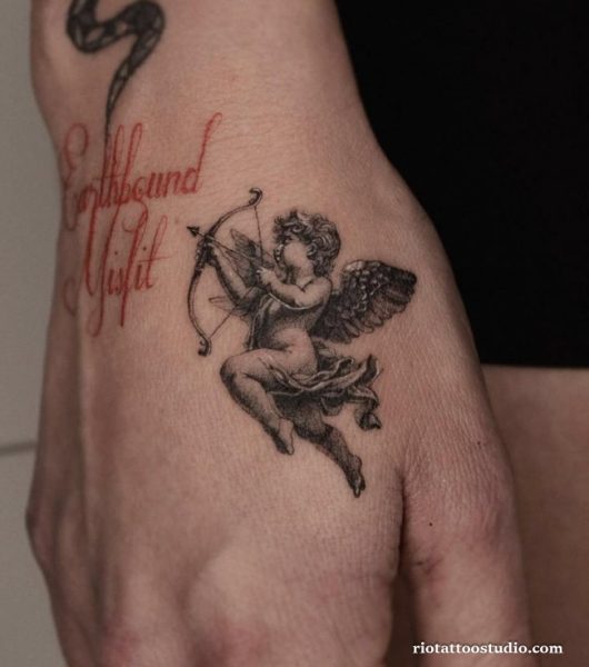 Cherub angel tattoo with bow and red script "Earthbound Misfit" on hand, unique cute angel tattoo