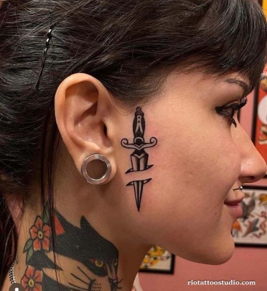 Dagger face tattoo with ornate handle on side face near ear, bold small face tattoo