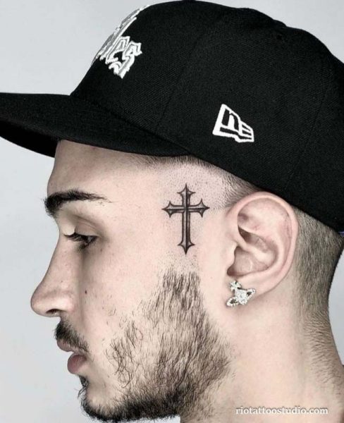 Ornate cross face tattoo on temple near ear, symbolic small face tattoo