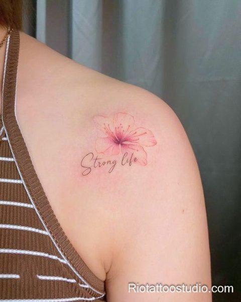 Pink hibiscus flower tattoo with "Strong life" script on shoulder, empowering feminine floral tattoo
