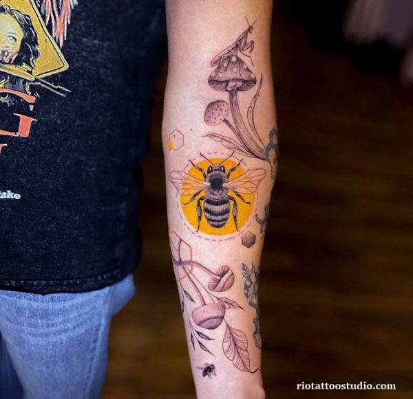 Bee tattoo with mushrooms, leaves and honeycomb in yellow circle on forearm