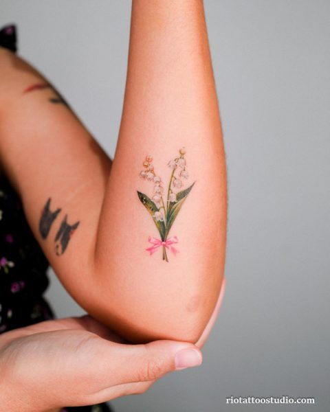 Minimalist birth flower tattoo with lily of the valley and pink bow on upper arm, delicate birth flower tattoo ideas