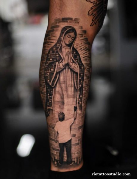 Chicano tattoo of Virgin Mary and child reaching for her on brick wall, realistic black and grey chicano tattoo on calf