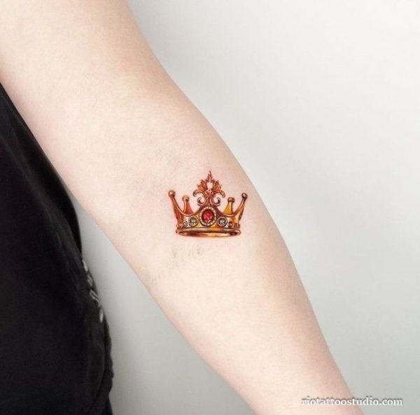 Small delicate golden crown tattoo with red jewel on forearm, minimalist crown tattoo ideas