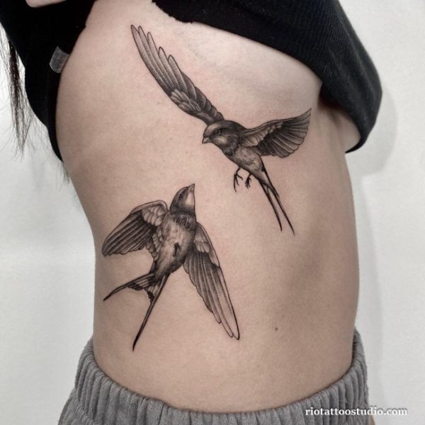 Delicate black and grey flying swallow birds rib tattoo on woman’s side, realistic bird tattoo ideas