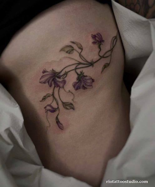 Watercolor purple flower vine rib tattoo on woman’s side, soft floral rib tattoo ideas