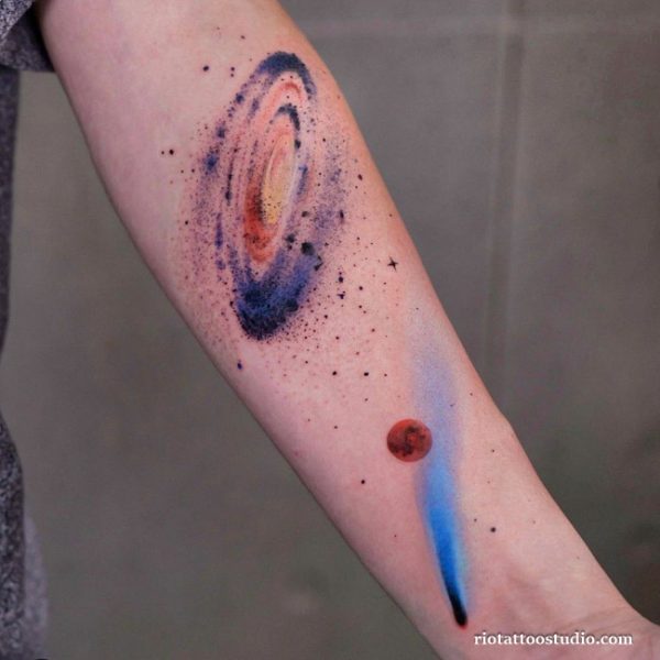 Unique watercolor tattoo on the forearm depicting a distant galaxy with deep purples and oranges, a red planet, and a blue comet trail with a watercolor gradient.