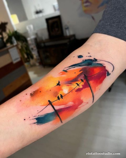 Vibrant watercolor sunset tattoo on the forearm featuring birds silhouette on power lines against an explosion of orange, red, and blue ink splatters.